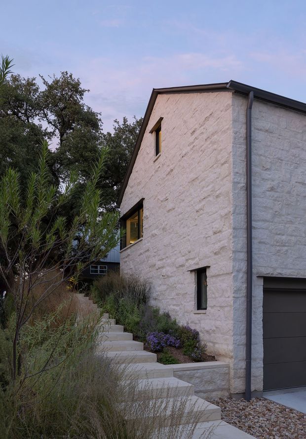 A vertical exterior view of a contemporary house with a steep gabled roof and pale rough-textured stone masonry walls. Narrow dark-framed windows and a dark garage door punctuate the facade, while concrete steps rise through a landscaped garden of grasses, shrubs, and trees. The muted evening light gives the scene a quiet, natural character.