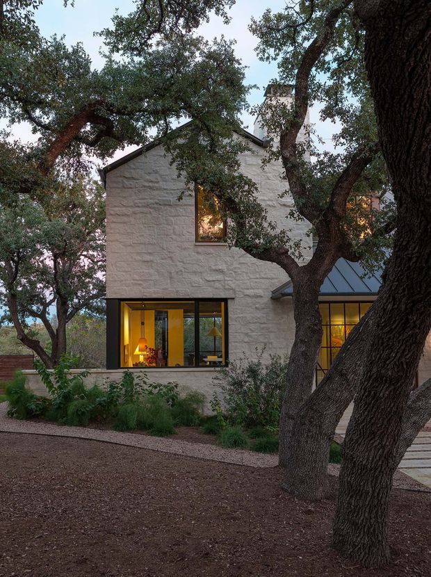 Exterior view of a contemporary multi-level home nestled among mature oak trees. The facade combines pale textured masonry with dark metal-framed glazing, a steep gabled roof, and a glazed lower-level extension. Warm interior lighting is visible through the windows, while native landscaping and a gravelled garden foreground reinforce the wooded setting.