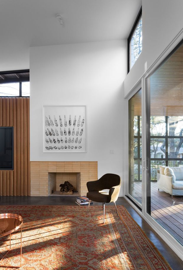 A bright, double-height living room with white walls and large floor-to-ceiling glazed doors opening onto a timber deck. The room features a warm light-brick fireplace, vertical timber wall panelling, dark timber flooring, a patterned red and orange area rug, and a sculptural brown lounge chair. A second seating area is visible through the glazing, with abundant natural light and leafy outdoor views.