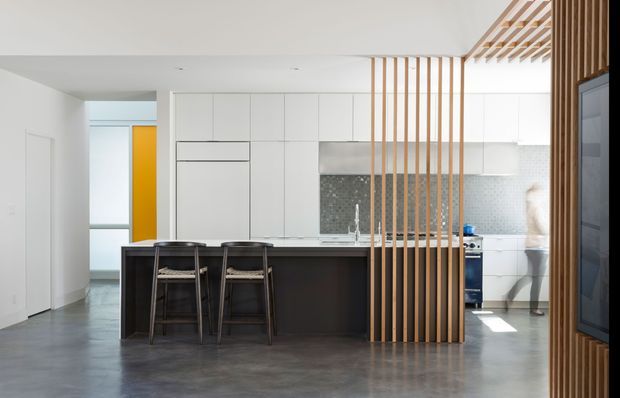 Bright contemporary open-plan kitchen viewed from the adjoining living area, with a large dark island and bar seating, white full-height cabinetry, a pale textured tile splashback, and a timber slat screen dividing the spaces. The polished grey concrete floor and white ceiling create a clean, minimal interior, with daylight entering through the rear glazing.