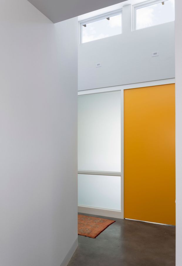 A minimalist interior hallway or entry area viewed from behind a partial wall. The space has white walls and ceiling, high clerestory windows, frosted-glass panels, a vivid mustard-orange full-height door, and a polished grey concrete floor. A small terracotta-toned rug adds warmth near the doorway. The high windows and vertical proportions suggest a tall, airy entry space.