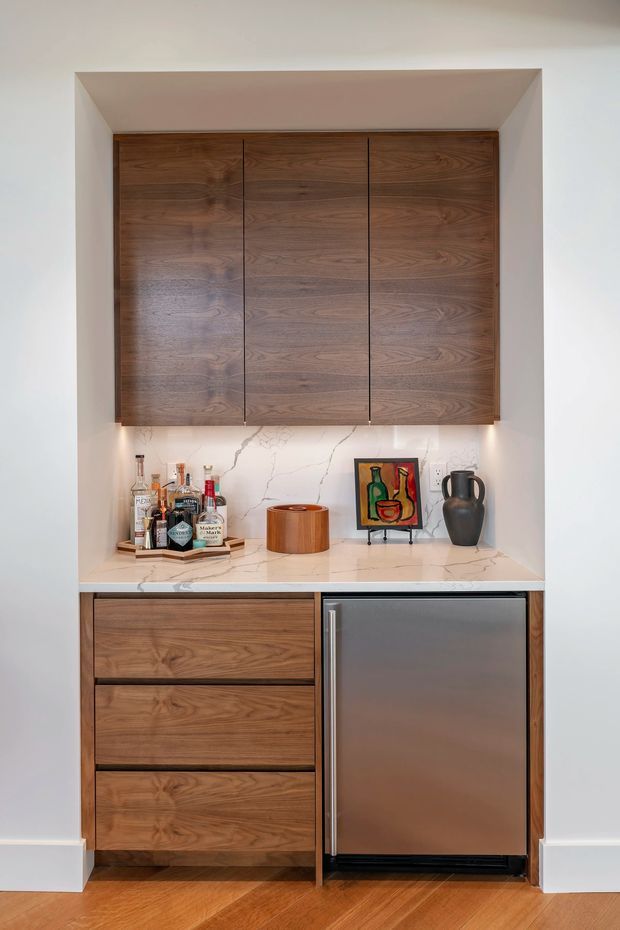 A compact built-in home bar or beverage station set into a white wall niche. The cabinetry has rich dark walnut-toned wood fronts with integrated vertical divisions above and drawers below, paired with a white marble-look stone benchtop and backsplash with subtle veining. A stainless-steel underbench refrigerator occupies the right side. Bottles, a small artwork, ceramic vessels and a bowl sit on the counter, with warm under-cabinet lighting and timber flooring continuing below.