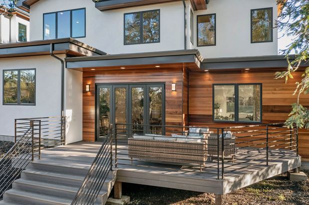 Exterior view of a contemporary multi-level home with white rendered walls, extensive dark-framed glazing, warm horizontal timber cladding, and a raised timber deck. Black metal railings, broad stairs, recessed soffit lighting, and a covered outdoor living area define the facade and entry.