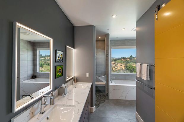 Contemporary bathroom with a long double vanity, white stone benchtop, wall-mounted mirrors with integrated lighting, grey tiled walls and floor, a freestanding bathtub visible through the glass shower enclosure, and a bold mustard-yellow door or cabinetry panel. Large windows bring in natural light and views of the surrounding landscape.