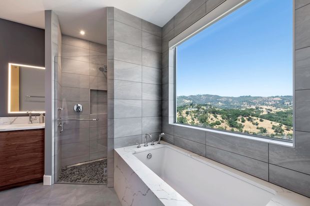 Contemporary bathroom featuring a freestanding-style built-in white bathtub beside a large picture window, a fully tiled glass-enclosed shower, double vanity with dark timber cabinetry, and an illuminated wall mirror. The grey large-format wall and floor tiles create a clean, refined finish, while the expansive mountain landscape is visible through the window.