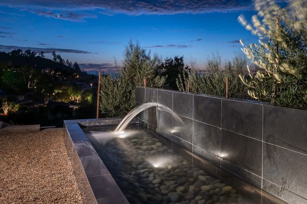 Twilight exterior view of a narrow linear water feature or reflecting pool, with illuminated smooth stones, a tiled feature wall, and a small cascade fountain. Dense planting and a distant suburban landscape surround the space, while gravel paving and subtle underwater lighting create a tranquil garden atmosphere.