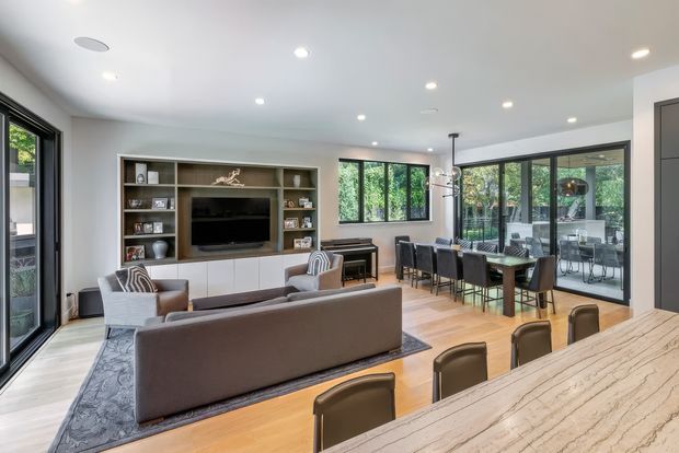 Open-plan contemporary living and dining area with a warm timber floor, large black-framed glazed doors and windows, and abundant recessed ceiling lighting. The living zone includes a sofa, armchairs, patterned rug, and built-in media cabinetry with shelving. A dining table and chairs sit beside the glazing, while a large stone-look island or countertop occupies the foreground. The space feels bright, spacious, and connected to greenery outside.