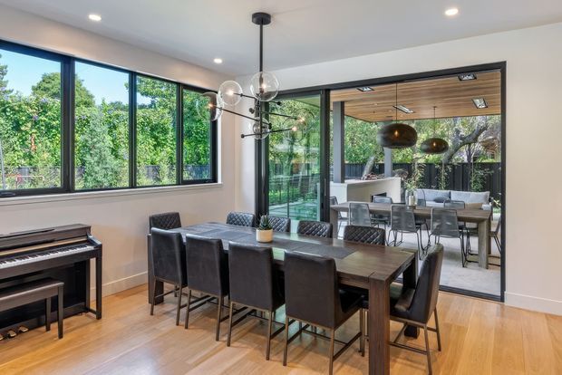 A contemporary dining room with a long dark timber dining table and upholstered dining chairs, opening through large black-framed glass doors to an outdoor entertaining area. Warm timber flooring, a black upright piano, sculptural pendant lighting, recessed ceiling lights, and extensive glazing create a refined indoor-outdoor setting.