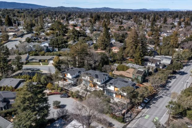 Aerial view of a suburban residential neighbourhood, showing a prominent multi-unit residential development along a broad tree-lined street, surrounded by detached homes, mature trees, local roads, and distant rolling hills.