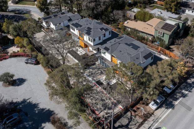 Aerial view of a contemporary multi-building residential development on a wooded suburban site. Several attached or closely grouped homes have dark pitched roofs, light exterior walls, large windows, and warm timber-clad accent sections. The homes are arranged along landscaped paths and driveways, with neighbouring houses and streets visible around the property.