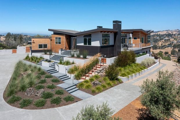 A low-profile contemporary single-storey residence on a dry, sloping site, with timber-clad walls, dark rendered wall sections, broad roof overhangs, a prominent dark chimney, and large windows. Terraced concrete paths and steps lead through a drought-tolerant landscaped garden with ornamental grasses, shrubs, gravel, and raised retaining beds. The surrounding arid hills are visible in the background.