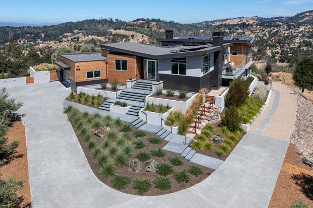 A modern multi-level hillside residence viewed from above, featuring dark grey and warm timber-clad exterior walls, broad low-slope roof forms, extensive outdoor stairs and terraces, concrete driveways and paths, and drought-tolerant landscaped planting. The home is set on a steep, rocky suburban or semi-rural site with expansive rolling hills visible in the background.