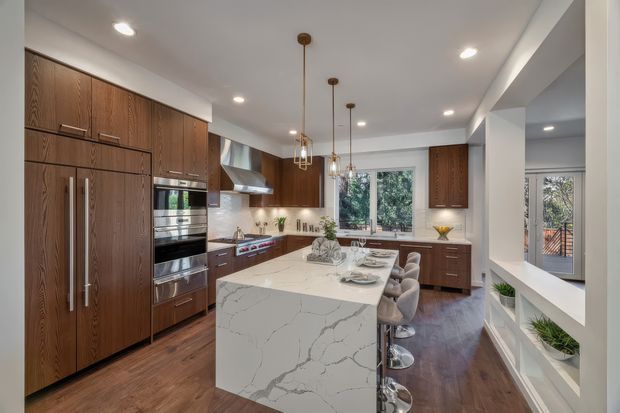 A spacious contemporary kitchen with full-height dark timber cabinetry, integrated stainless-steel appliances, a large white veined stone island with bar seating, pendant lights, recessed ceiling lighting, and dark timber flooring. A dining or living area is visible beyond the kitchen, with large windows overlooking greenery.