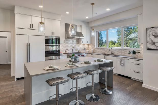 Bright contemporary kitchen with a large central island and seating for four. The space features white flat-panel cabinetry, integrated stainless-steel appliances, a pale stone-look benchtop, a white geometric tile splashback, timber-look flooring, pendant lighting, and broad windows overlooking greenery.