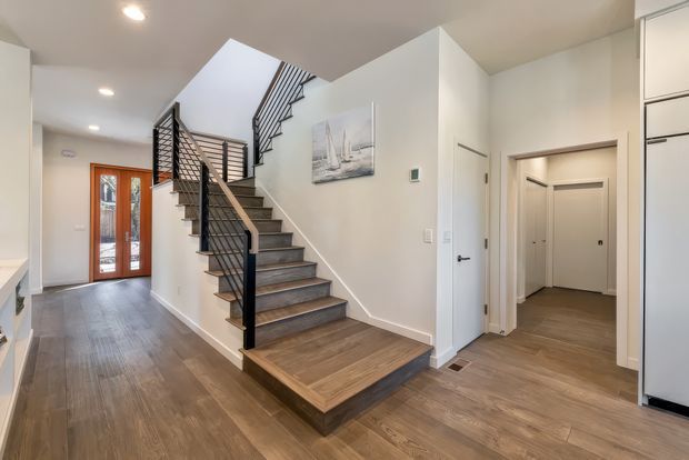 Bright contemporary residential entry hall with a broad timber-look flooring, a statement open-riser staircase with dark metal balustrade and timber treads, white walls, recessed ceiling lights, built-in cabinetry, and glazed timber double entry doors. A corridor and interior doors lead to adjoining rooms.