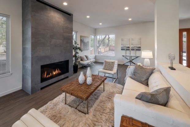 Bright contemporary living room with a large grey stone-clad fireplace wall and linear fireplace, dark timber flooring, white sectional seating, textured neutral rug, and a rustic timber coffee table. Large windows bring in natural light, while recessed ceiling lights and neutral artwork create a calm, refined atmosphere. A dining or adjacent space is partially visible beyond the opening.
