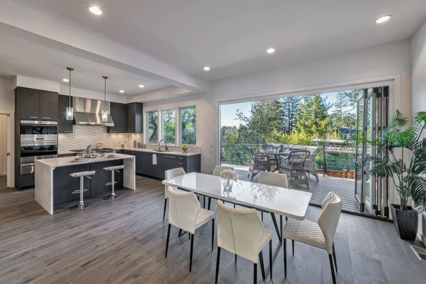 Bright open-plan kitchen and dining area with dark charcoal cabinetry, a light stone island and benchtops, and a white subway-tile splashback. A glossy dining table sits beside floor-to-ceiling sliding glass doors opening to a timber deck, with abundant greenery visible outside. Recessed ceiling lighting, pendant lights, and grey-toned timber flooring create a contemporary, airy interior.