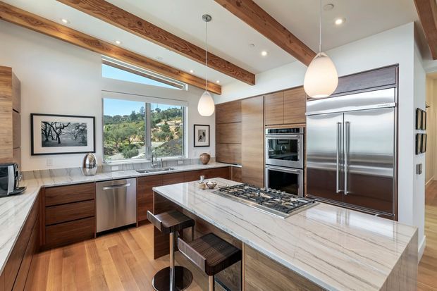 A spacious, refined kitchen with a large marble-look island and integrated gas cooktop, bar seating, warm timber cabinetry, stainless-steel appliances, and a window overlooking a leafy landscape. Exposed timber ceiling beams and pendant lights add warmth and character.