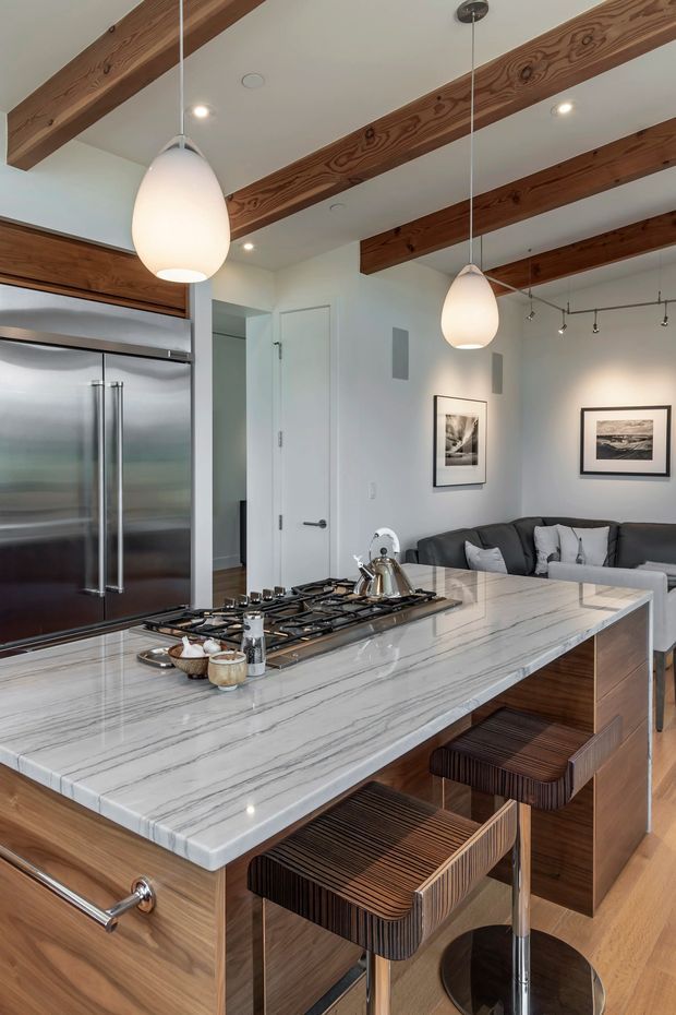 A contemporary open-plan kitchen with a large waterfall-edge marble island, integrated gas cooktop, timber cabinetry, stainless-steel refrigerator, and two timber bar stools. Exposed timber ceiling beams, white walls, pendant lights, and recessed lighting define the space, with a living area and sofa visible beyond the island.