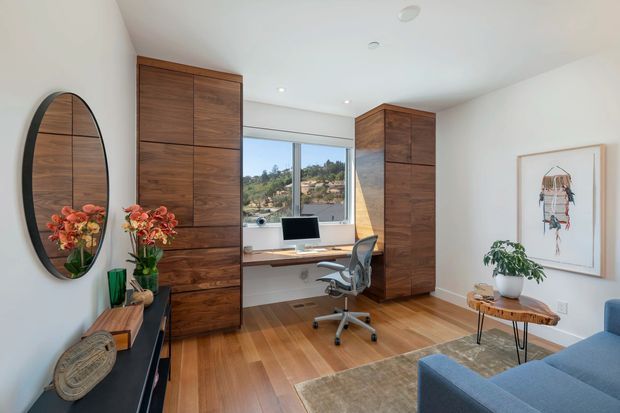 Bright, refined home office with extensive dark timber built-in cabinetry flanking a window, a compact integrated desk and ergonomic office chair, warm timber flooring, and white walls. The room includes a large oval mirror, framed artwork, decorative plants, a console, rug, and part of a blue sofa. A leafy residential view is visible through the window.
