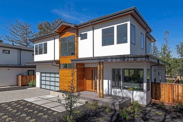 A contemporary two-storey residential house viewed from the front, with white rendered walls, dark trim, warm timber cladding around the entry and upper window bay, a low-pitched roof with broad eaves, large glazed windows, an attached garage, paved entry path, and timber fencing. The landscaped front garden and clear blue sky surround the modern facade.