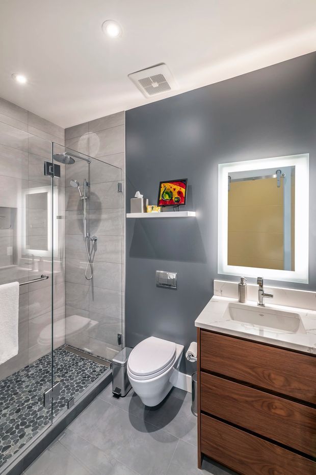 Contemporary bathroom featuring a frameless glass walk-in shower with grey wall tiles and pebble mosaic shower flooring, a wall-mounted toilet, floating dark timber vanity with white countertop and integrated basin, and a large illuminated mirror. Recessed ceiling lights and a ceiling exhaust fan provide bright, clean illumination.