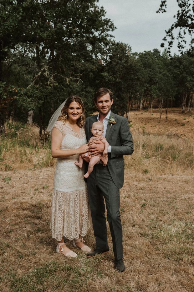 A portrait-oriented outdoor photograph of a bride and groom standing together in a dry grassy clearing, holding a baby. The bride wears a lace wedding dress and veil, while the groom wears a dark tailored suit with a boutonniere. Mature trees and woodland form the background, creating a warm, rustic wedding setting. No building or architectural space is visible.