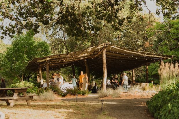 A rustic open-air timber pavilion set within a leafy garden, with a rough natural pole structure and layered branch-like roof covering. People gather around tables beneath the shelter, while ornamental grasses, mature trees and informal landscaping surround the space. The scene has a warm, natural outdoor-event atmosphere.