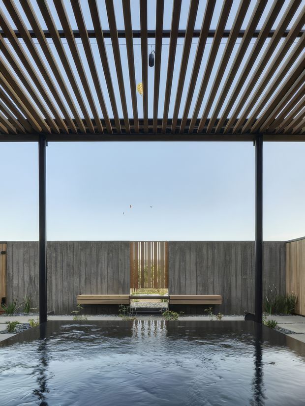 A tranquil outdoor courtyard with a shallow reflecting or swimming pool in the foreground, enclosed by dark timber privacy fencing and a central slatted timber gate. A covered pergola with closely spaced timber battens and slim dark structural posts shelters the space, with built-in bench seating and sparse architectural planting around the pool. The composition is minimalist and strongly geometric, with open sky visible through the pergola.