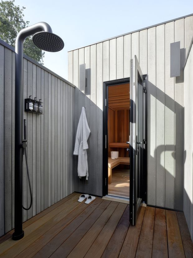 A modern outdoor shower courtyard with dark timber decking, grey vertically panelled walls, and a freestanding black rainfall shower. An open glazed door leads into a compact timber-lined sauna with built-in bench seating. A robe, sandals, and shower products reinforce the spa-like use of the space.