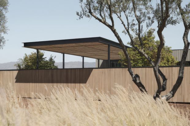 A modern low-profile outdoor structure with a broad flat roof and exposed timber soffit, supported by slender dark metal posts. It sits behind a tall vertical timber privacy fence, with muted desert landscaping and mature trees in the foreground and distant rugged hills beyond. The composition emphasizes the building’s horizontal lines, warm timber tones, and integration with the dry natural setting.