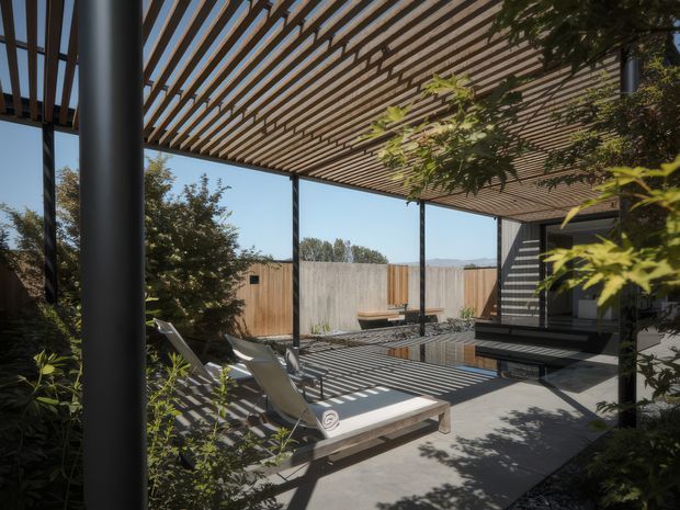A contemporary outdoor patio or terrace beneath a large timber-slatted pergola. Slim dark structural posts support the open roof, casting strong linear shadows across a concrete-paved entertaining area. The space includes reclining lounge chairs, a low timber bench, a dark built-in outdoor kitchen or barbecue zone, planted borders, and a timber privacy fence. The setting is sunny, private, and landscaped, with a restrained palette of dark grey, natural timber, and greenery.