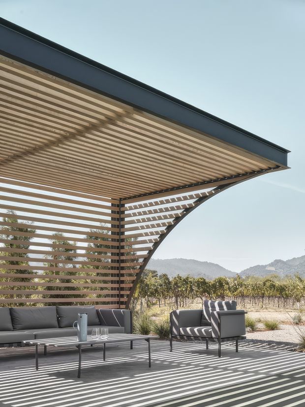Outdoor lounge beneath a dramatic cantilevered pergola with a curved slatted timber privacy screen and matching slatted ceiling. The space includes a dark grey sectional sofa, armchairs, and a low coffee table, opening toward a vineyard and mountainous landscape. Strong linear shadows create a graphic, contemporary atmosphere.