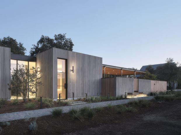 Twilight exterior view of a contemporary, low-rise building composed of several rectilinear volumes clad in vertically ribbed grey metal or timber-like panels. Floor-to-ceiling glazed openings reveal warmly lit interiors, while a covered open terrace with a timber-toned ceiling connects to a lower wing. A gravel path and restrained native-style landscaping occupy the foreground.