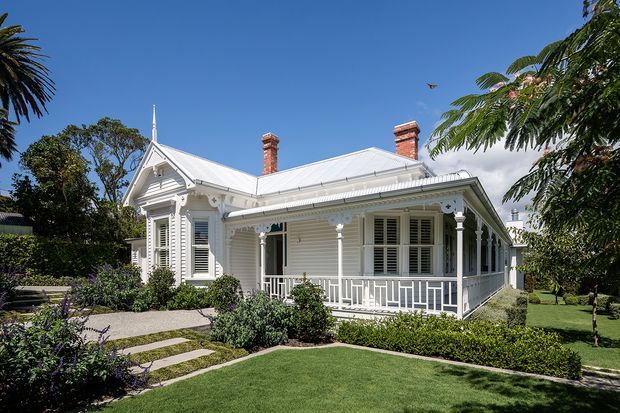 A well-maintained single-storey historic timber villa with a pale weatherboard facade, decorative white trims, a steep hipped and gabled metal roof, twin brick chimneys, ornate verandah detailing, louvred shutters, and a prominent front porch. The house is set within a manicured garden with clipped hedges, lawn, stepping-stone paving, and mature tropical vegetation under a clear blue sky.