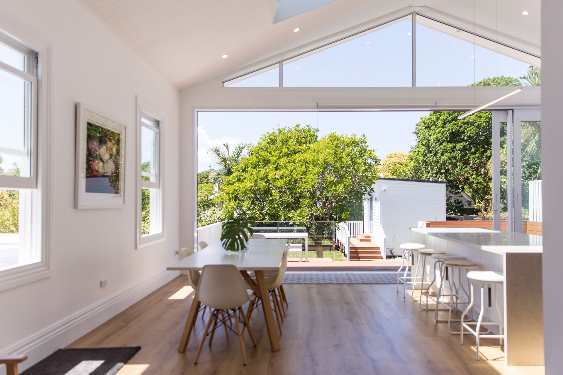Full-width stacking sliders dissolving the boundary between dining room and deck — with the separately designed garden studio quietly visible beyond. Grey Lynn Villa, Macfie Architecture.