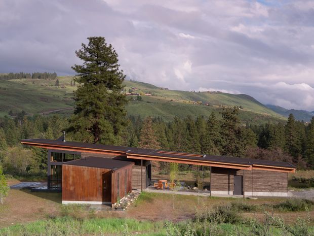 A low-slung contemporary house set in a mountainous, forested landscape. The single-storey building has an elongated flat or very low-pitched roof with broad overhangs, dark horizontal cladding, extensive glazing, and a prominent weathered timber-clad box-like volume at the front. Rolling green hills, coniferous trees, and dramatic clouds form the background.