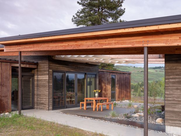 A single-storey modern timber-clad building with a broad covered outdoor dining area. The structure features exposed timber roof beams, slender dark metal posts, a low-slope dark roof, large glazed doors, and a simple orange timber dining table and benches. Gravel landscaping and rolling rural hills are visible beyond the courtyard-like covered space.