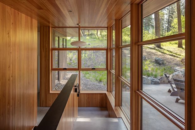 A narrow, light-filled interior passage or living space lined with warm timber wall and ceiling panelling. Floor-to-ceiling timber-framed glazing provides views to a wooded, rocky landscape and an outdoor seating area. A dark linear built-in element extends through the space, while a simple pendant light and polished concrete floor contribute to the restrained modern design.