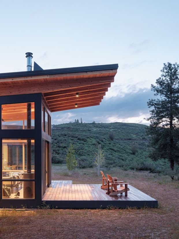A compact modern cabin viewed from outside at dusk, set in a rugged, shrub-covered landscape. The building has a dark minimalist exterior, expansive floor-to-ceiling glazing, a broad low-pitched roof with exposed timber eaves and recessed lights, and a timber deck furnished with two wooden Adirondack-style chairs. Warm interior lighting reveals a small living area through the windows. A metal chimney rises from the roof.