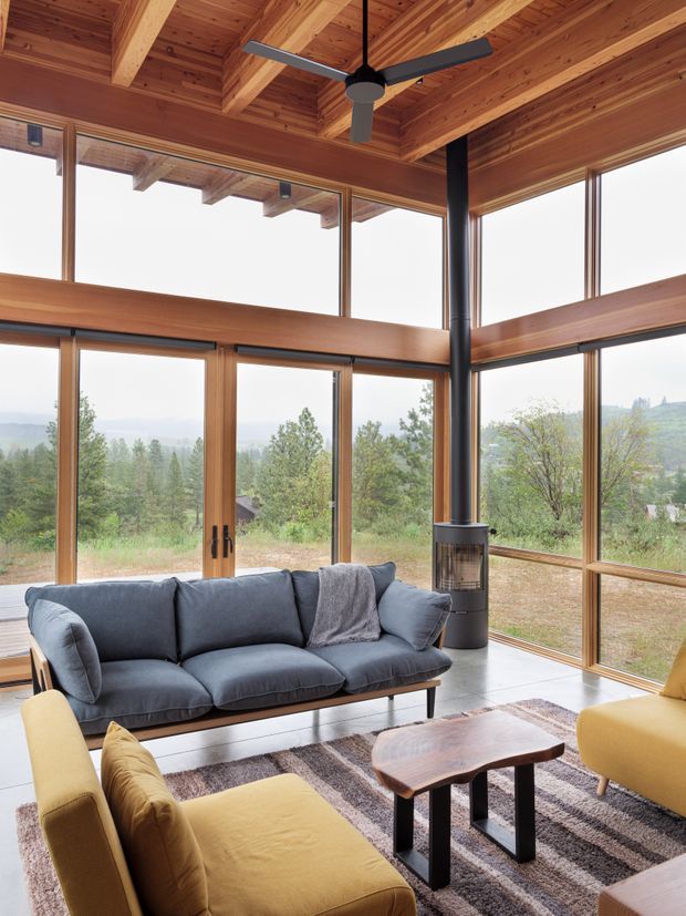 A bright, timber-lined living room with extensive floor-to-ceiling glazing overlooking a wooded landscape. The room features a dark freestanding wood-burning stove with flue, exposed timber ceiling beams, a ceiling fan, a deep blue-grey sofa, mustard lounge chairs, and a low live-edge timber coffee table on a patterned rug.