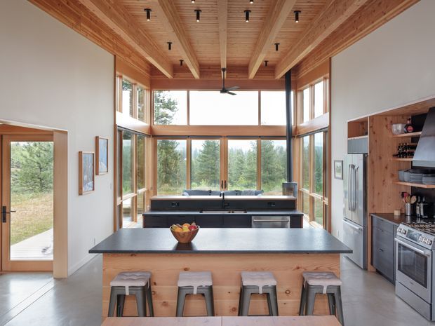 A spacious contemporary kitchen with a large central island, dark stone benchtop, timber-panelled island base, and four stools. The room features extensive floor-to-ceiling timber-framed glazing overlooking a wooded landscape, exposed timber ceiling beams and boards, integrated ceiling spotlights, and a ceiling fan. Built-in cabinetry, stainless-steel refrigerator and range cooker line the side wall, while a secondary sink bench sits in front of the windows.