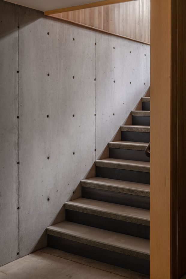 A narrow interior stairway with concrete walls featuring exposed form-tie holes, dark stair risers, and light stone or concrete treads. Warm timber wall panelling and a timber door or frame add contrast, creating a restrained, material-focused modern interior.
