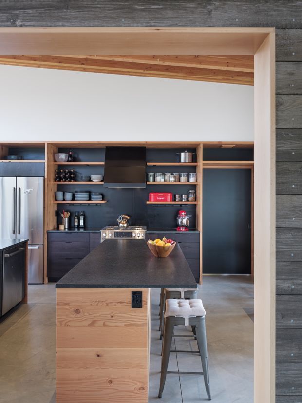 A contemporary kitchen viewed through a broad timber-framed opening. The room features a large dark stone island with timber panelled ends, integrated seating, dark lower cabinetry, open timber shelving, a dark rangehood, stainless-steel appliances, and a polished concrete floor. Timber ceiling detailing and dark horizontal exterior-style wall cladding frame the space.