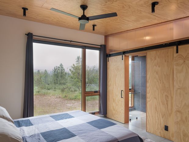 A compact, warmly finished bedroom with plywood wall and ceiling linings, a large glazed window overlooking vegetation, dark curtains, and a patchwork bedspread. A ceiling fan and recessed spotlights provide lighting and ventilation. Sliding plywood doors open to a tiled bathroom or adjoining wet area, while the room has a simple contemporary cabin-like character.