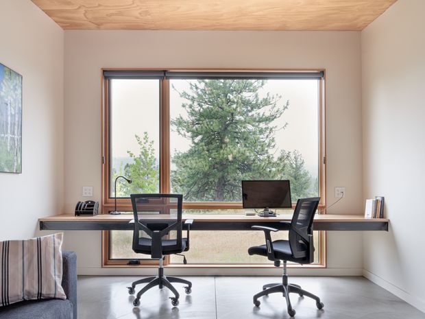 A bright, compact home office with two black ergonomic swivel chairs positioned at a long timber desk spanning a large window. The room has pale walls, a warm timber-lined ceiling, a polished light concrete floor, timber-framed glazing, and views of trees outside. Minimal office equipment and shelving create a clean, functional workspace, with part of a sofa visible at the left edge.
