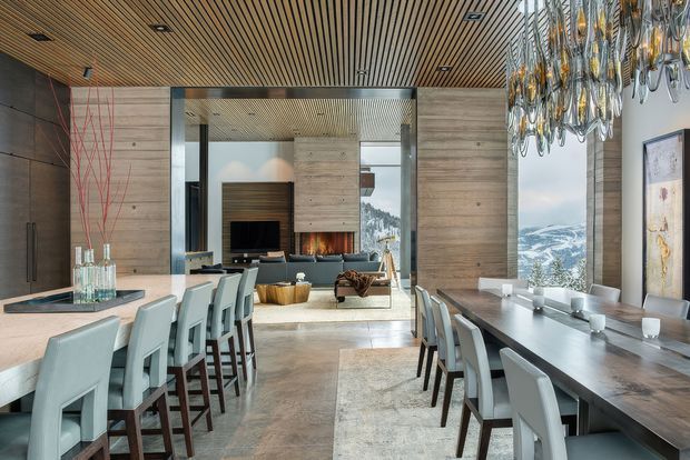 A sophisticated open-plan dining and living area with a long dark dining table, pale upholstered dining chairs, and a kitchen island with bar seating in the foreground. Warm timber slatted ceilings, large-format timber wall panelling, polished concrete flooring, and a sculptural clustered pendant light define the interior. Beyond the dining area is a lounge with a television and fireplace, while expansive glazing reveals a dramatic snow-covered mountain landscape.