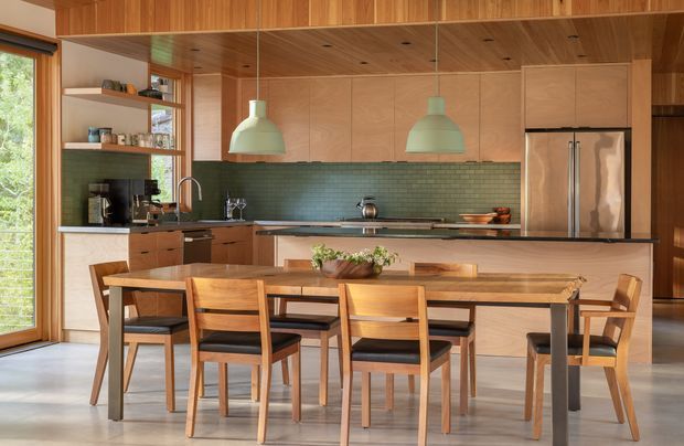 Warm contemporary open-plan kitchen and dining area with extensive timber wall and ceiling lining, integrated timber cabinetry, a dark green tiled splashback, pale stone island benchtop, stainless-steel refrigerator, and a long solid timber dining table with matching timber chairs. Large glazing on the left brings in natural light and greenery views.