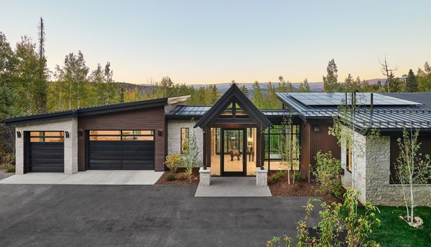 Contemporary single-storey residential building viewed from the front, with a low-pitched intersecting roofline, dark metal roofing, timber and light masonry cladding, a glazed gabled entry porch, and a two-car garage. The landscaped setting and distant wooded hills are visible behind the house.