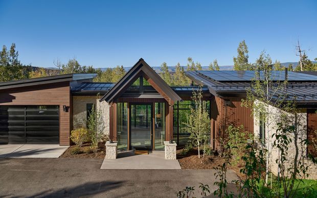 Exterior view of a low-slung contemporary lodge-style residence with a prominent timber-framed gabled entry, extensive dark-framed glazing, warm brown cladding, integrated garage, and broad dark metal roof forms. The house is set among landscaped trees with distant hills visible beyond.